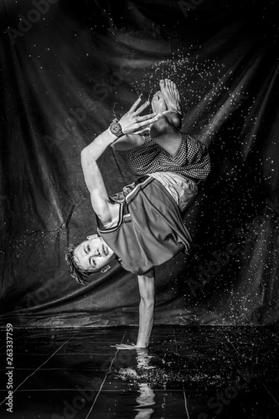 Fototapeta cool and happy korean young guy break dancer in style of bboying doing complex tricks on floor with water splashes and liquid drops effects in aqua zone photo studio with black isolated background