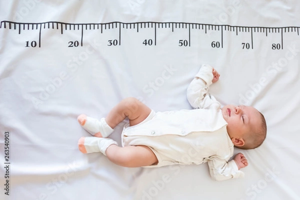 Fototapeta A newborn 2 month baby in white sleeps on a bed on which a measuring ruler for growth is drawn