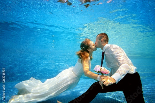 Obraz The beautiful bride and groom hold hands and kiss underwater at the bottom of the pool on a Sunny day. Portrait. Concept. Underwater wedding. Horizontal view