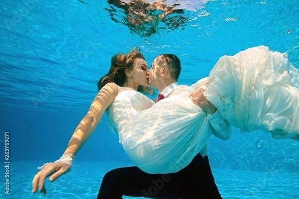Obraz Happy bride and groom kissing underwater in the pool on a Sunny day. The groom hugs and holds the bride in his arms at the bottom. Portrait. Concept. Wedding underwater. Horizontal view
