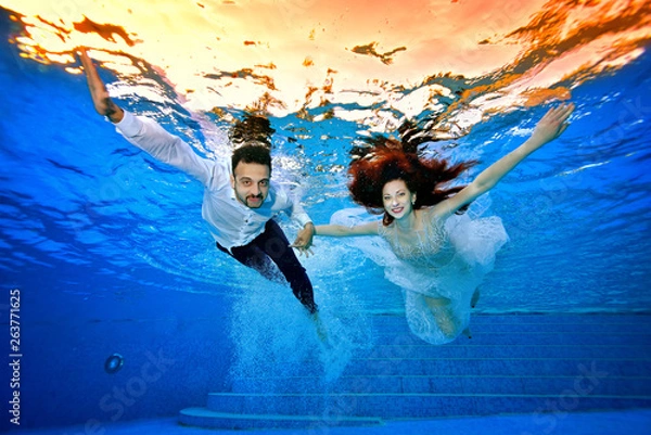 Obraz Happy bride and groom in wedding dresses swim underwater on the background of a bright sunset on the surface of the water. They smile and pose for the camera with their arms outstretched. Portrait