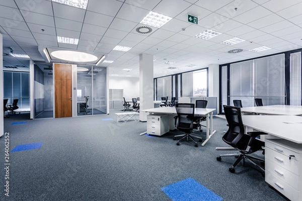 Fototapeta Part of ordinary office room decorated in modern style with meeting room,large windows with blinds,carpeting,ventilation,escape signs,white furniture (tables, shelves, drawers) and dark office chairs.