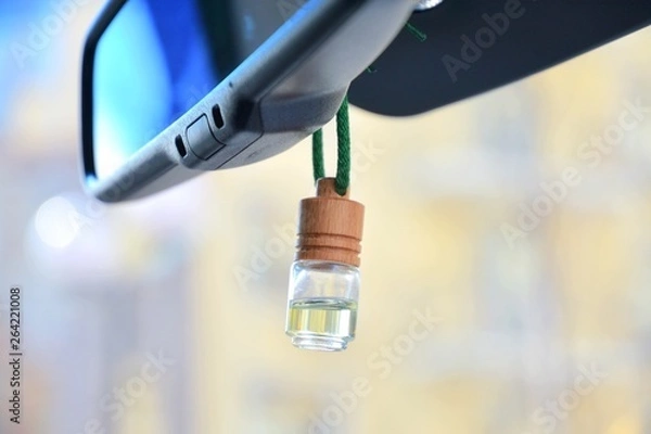 Obraz Car air perfume freshener bottle inside the car with part of car mirror on blurred city background. Little glass bottle with wooden lid and yellow aromatic liquid automobile freshener on a green rope.