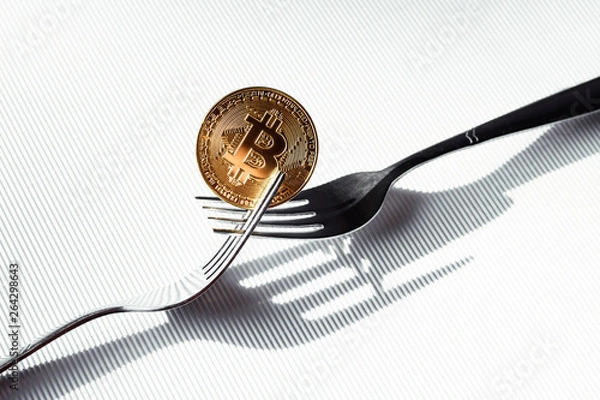 Obraz Shiny gold coin with bitcoin symbol between two silver forks on white background. Concept of virtual crypto currency.