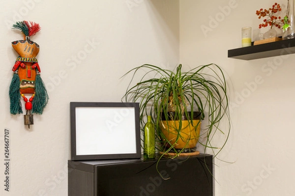 Fototapeta Blank black horizontal picture frame on a shelf with a ponytail palm  and a tribal mask. For mocking up photos and artwork.