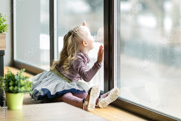 Fototapeta A beautiful child is sitting near a large window. A little girl, 4-5 years old, is sitting on the windowsill of a large window in  cafe.