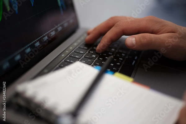 Fototapeta Close-up of man hands, typing on keyboard of laptop and holding mug of coffee, near notebook with black pencil.
