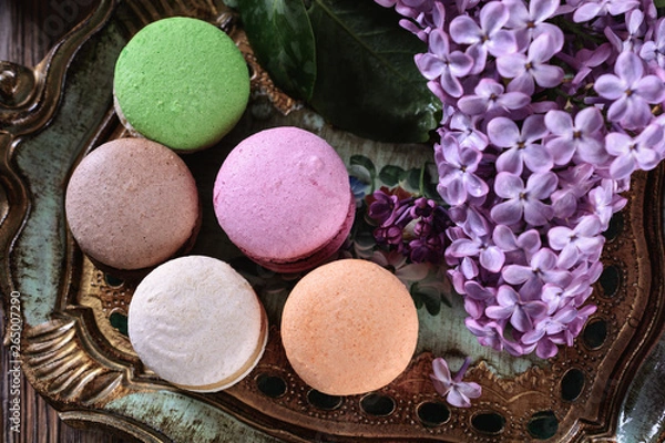 Fototapeta Multicolored bright macaroons on a wooden vintage tray next to flowers of lilac on a wooden table, top view