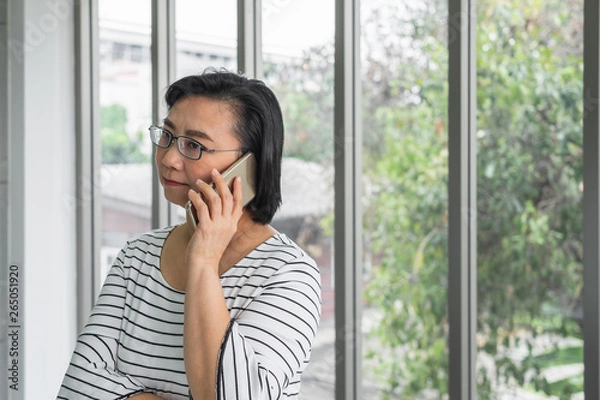 Obraz An old Asian woman mother holding a phone in front of a window looking concern and serious.