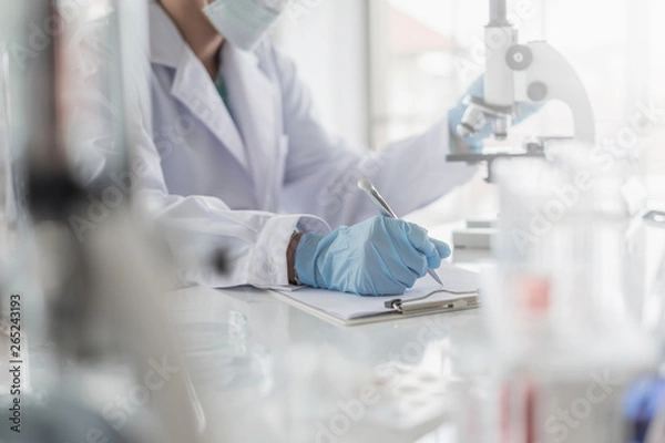 Obraz A scientist hands writing on a clipboard in laboratory with test tube microscope and solutions.