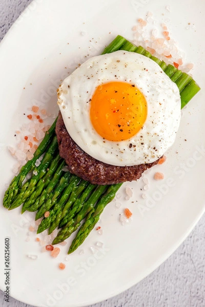 Fototapeta Beefsteak from minced beef with fried eggs and fresh green asparagus. Ketogenic food. Large portion on a white plate. Close up and vertical view.