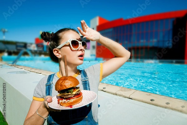 Fototapeta A girl with a Burger by the pool, a model in sunglasses holding a Burger. The concept of juicy burgers.