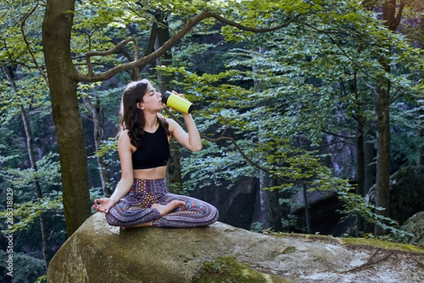 Fototapeta Healthy fitness girl sitting in lotus pose and drinking water from green bottle in forest