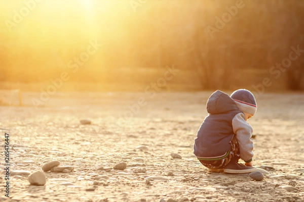 Fototapeta A six-year-old boy with autism sits on the ground alone at sunset. Lonely little child playing with stones outside. The concept of autism and loneliness. Copy space for your text. Setting sun.