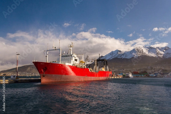 Obraz Red container ship in Ushuaia port. A big colorful cargo ship on beagle channel on a sunny winter day with the city and mountains on the background