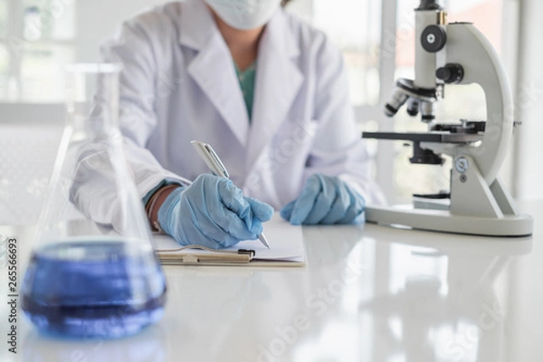 Obraz A scientist hands writing on a clipboard in laboratory with test tube microscope and solutions.