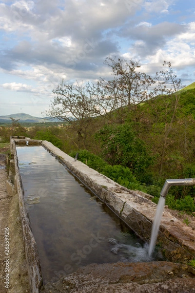 Fototapeta Source of spring water in the forest, small mountain village in the background.