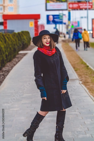 Fototapeta The happy girl with smile walks around the city in black coat and hat with wide fields. Red lipstick and scarf. Fashion, style.