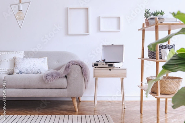 Fototapeta Stylish and cozy scandinavian white interior with design sofa, bookstand with accessories, pillows, blanket, gramophone and mock up photo frames. White background walls and modern triangle lamp. 