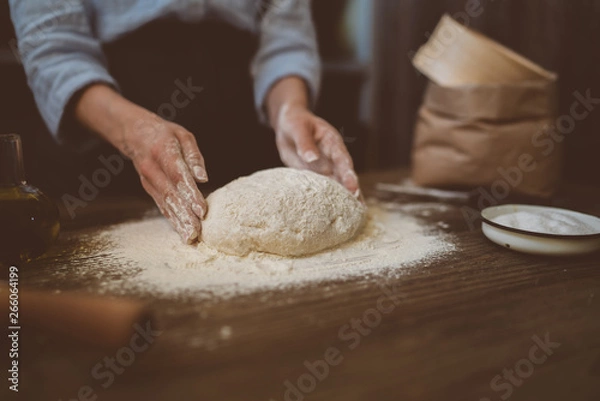 Fototapeta Female hands with dough in ball shape. Basic homemade dough with ingredients on wooden table with natural light. Home healthy food. Low key shot, close up on hands, some ingredients around on table.