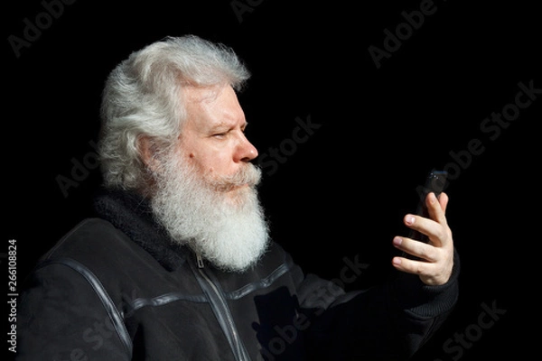 Fototapeta Handsome senior man looking at smartphone against black background. Close up profile portrait of elderly stylish male with splendid mustache and beard wearing dark flight (bomber) jacket.