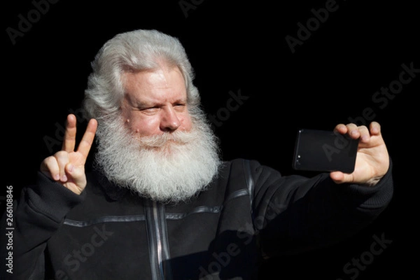 Fototapeta Handsome senior man looking at smartphone and showing a peace sign against nature background. Close up face portrait of elderly stylish male with splendid mustache and beard wearing dark flight jacket