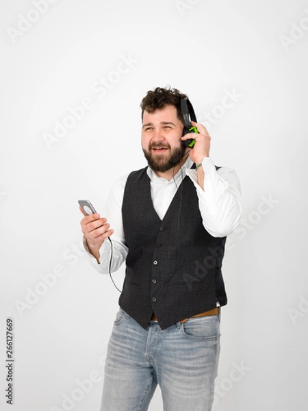 Fototapeta cool man with black beard and white shirt is posing with headphones and smartphone in front of white background