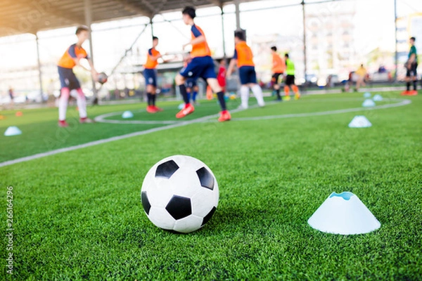 Fototapeta football between marker cones on green artificial turf with blurry soccer team training