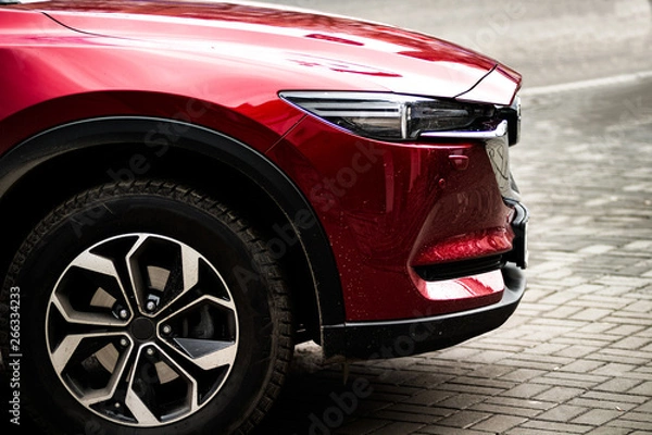 Fototapeta The front side of a red car with a large bright wheel standing on a wet asphalt. Background.