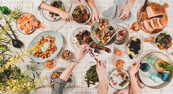 Fototapeta Family or friends gathering dinner. Flat-lay of peoples hands, roasted lamb shoulder, salads, vegetables, rose wine, mimosa branches over white checkered tablecloth, top view. Celebration party dinner