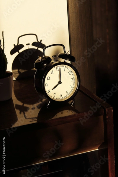 Fototapeta Black retro vintage alarm clock on a wooden bedside table with reflection in a polished surface in the rays of the setting sun with hard shadows