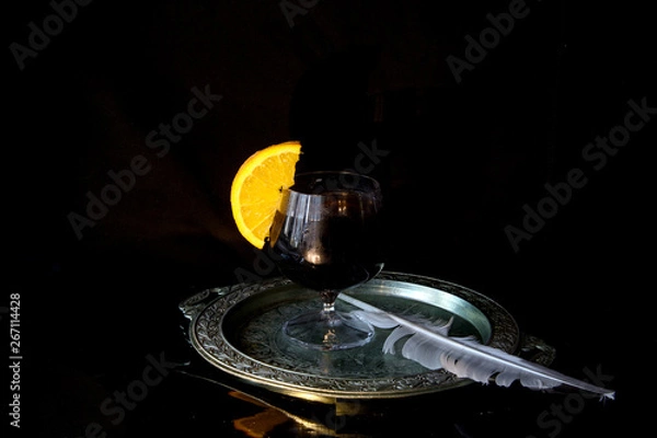 Obraz On a silver tray is a glass of red wine with an orange slice on the edge. Nearby is a goose feather. Concept - a mysterious evening