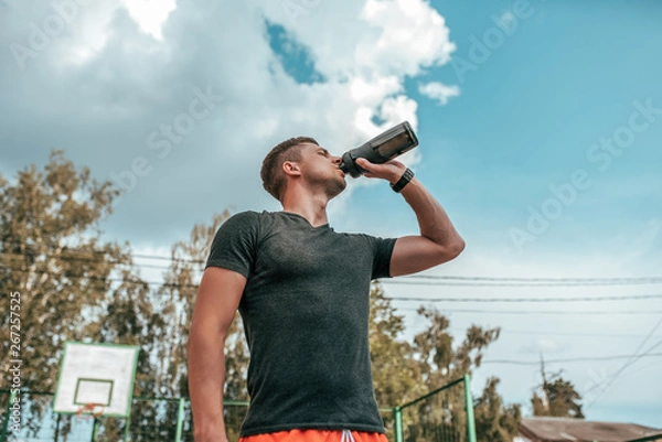Fototapeta Male athlete, drinking water and protein from shaker, fitness watch, training in the city in the fresh air. Concept morning run in the city, fitness and workout, smart watch bracelet. Sportswear T