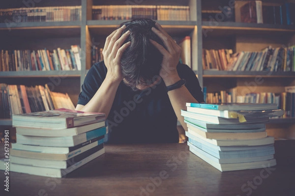 Fototapeta Stress and tired man under mental pressure while reading book preparing examination in library.