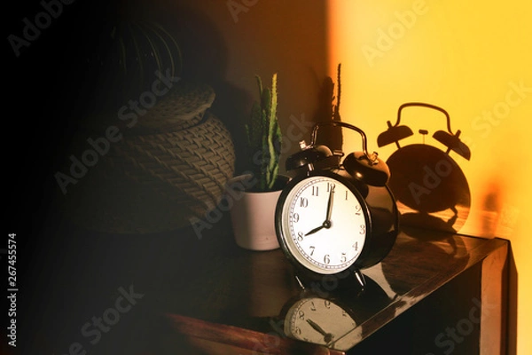 Fototapeta Black retro vintage alarm watch clock on a wooden bedside table with reflection in a polished surface in the rays of the setting sun with hard shadows. Sepia toning