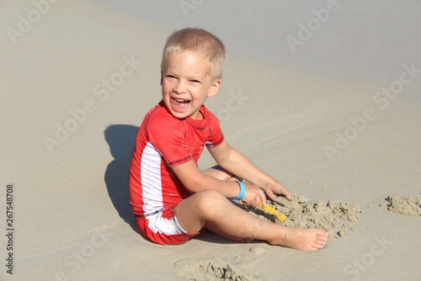 Fototapeta A little blond boy in clothes with UV filter is playing with sand on the beach by the sea, holiday with children, protecting kids from the sun concept