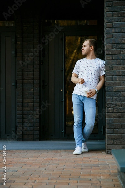 Fototapeta A man stands on the porch of a brick building and drinks coffee from a paper Cup in the morning