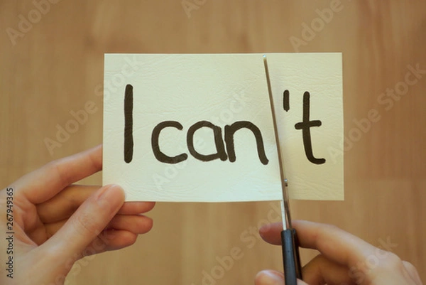 Obraz I can, I can't. Hand holding white paper and scissors in front of brown background with a message I can