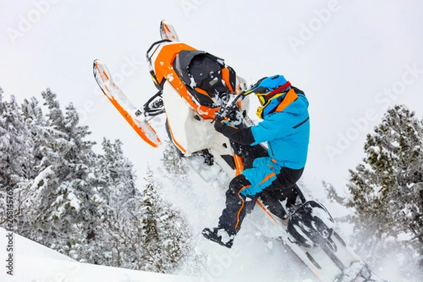 Obraz snowmobile breaks out of the snow. close-up of a sports snowmobile with a pilot on a background of gray snow-covered forest. bright snowmobile and suit without brands. extra high quality
