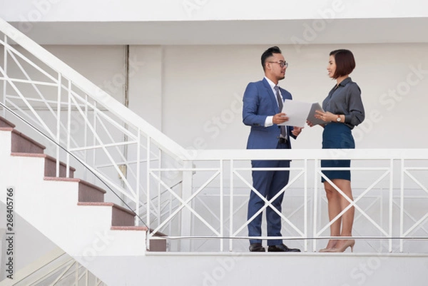 Fototapeta Two young business people holding document and tablet pc talking to each other while standing on staircase at office