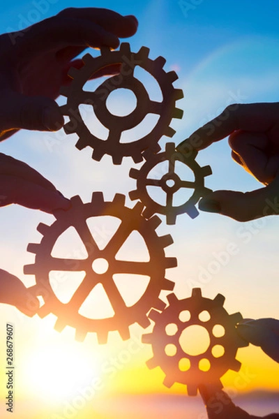 Fototapeta Gears in the hands of people on the background of the evening sky. the mechanism of teamwork. interaction.