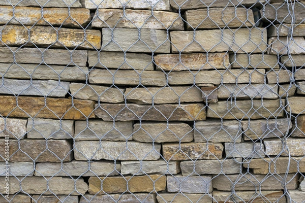 Fototapeta The construction of the wall of natural stone. The stone slabs are stacked on top of each other. A pile of natural stone is fenced with a metal mesh.