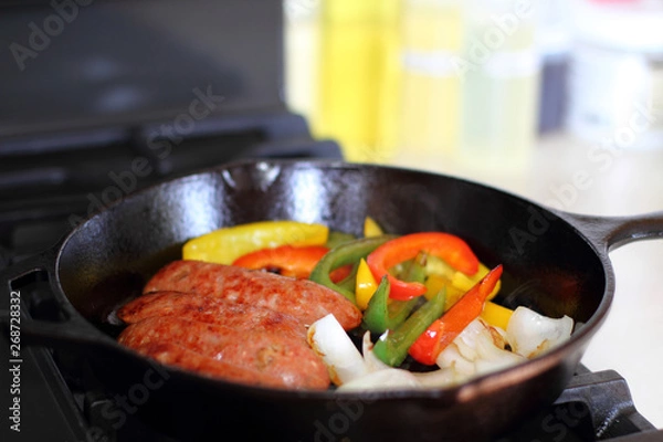 Fototapeta Sausage cooking on the stove in a cast iron skillet with bell peppers and onion.