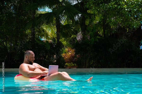 Fototapeta male with an inflatable ring in the water in the pool works on the computer. Funny Fat Man in Swimming Circle with Pink Laptop. freelance concept