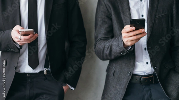 Fototapeta Social media addiction. Modern corporate life break time. Two men in business suits leaning on the wall, using smartphones.
