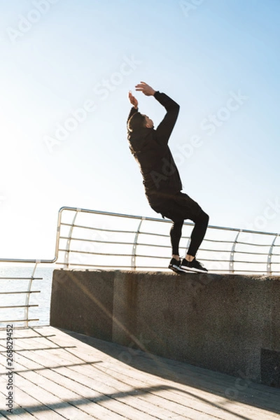 Fototapeta Photo of strong guy 20s doing acrobatics and jumping during morning workout by seaside