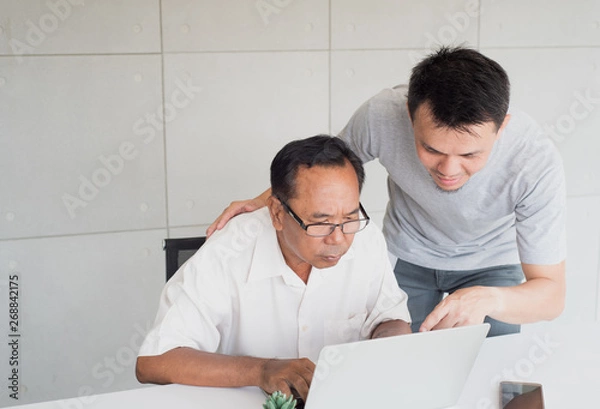 Fototapeta Young asian man in casual shirt with feeling confidence teach senior asian man with feeling confuse to use computer notebook in modern office background, family activity, copy space on top left