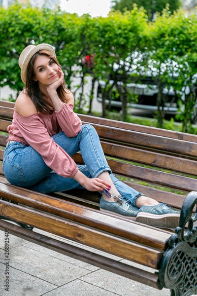 Obraz Fashionably dressed young pretty woman on the street on a sunny summer evening. Beautiful girl in jeans, a blouse, small hat and sunglass is sitting on a bench and smiling. Street-fashion concept