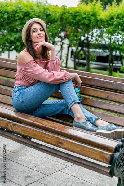 Obraz Fashionably dressed young pretty woman on the street on a sunny summer evening. Beautiful girl in jeans, a blouse, small hat and sunglass is sitting on a bench and smiling. Street-fashion concept