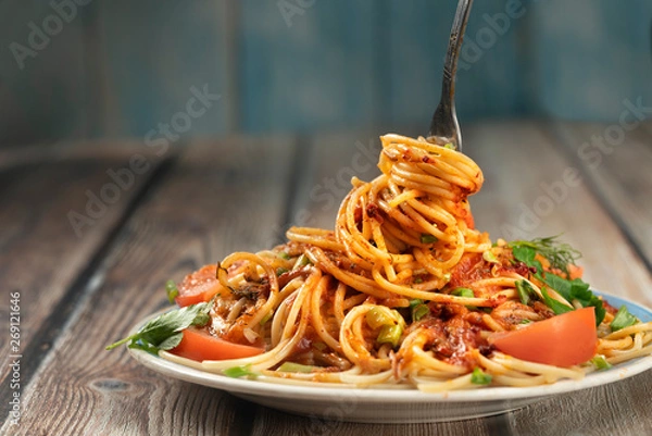Fototapeta plate of pasta seasoned with herbs, tomatoes and sauce on a wooden table close-up, fork with long pasta and sauce, share menu restaurant and cafe